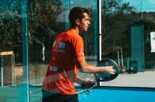 Joueur derrière la paroi vitrée d'un court de padel, surface bleue et fixations visibles